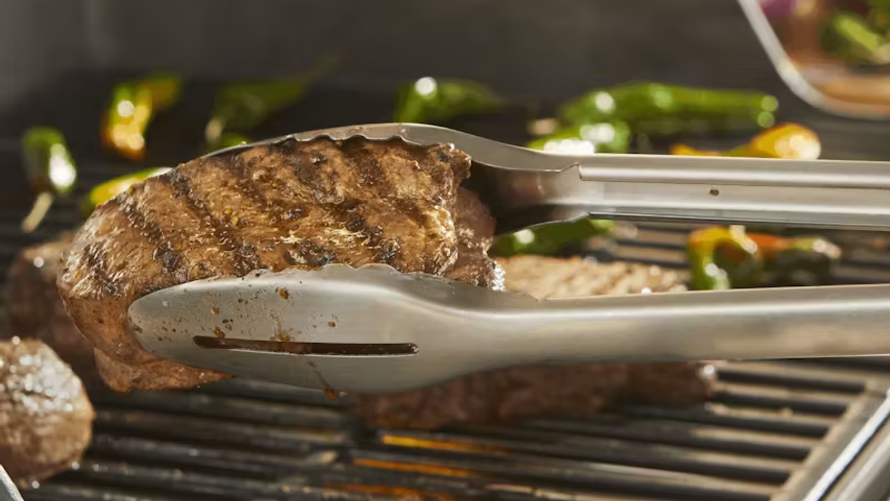 En grilltang holder et stykke grillet kød over en varm grill med peberfrugter.